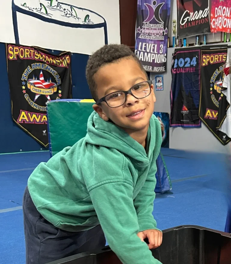 A smiling young boy wearing glasses and a green hooded sweatshirt leans forward in a gym setting with cheerleading banners in the background.