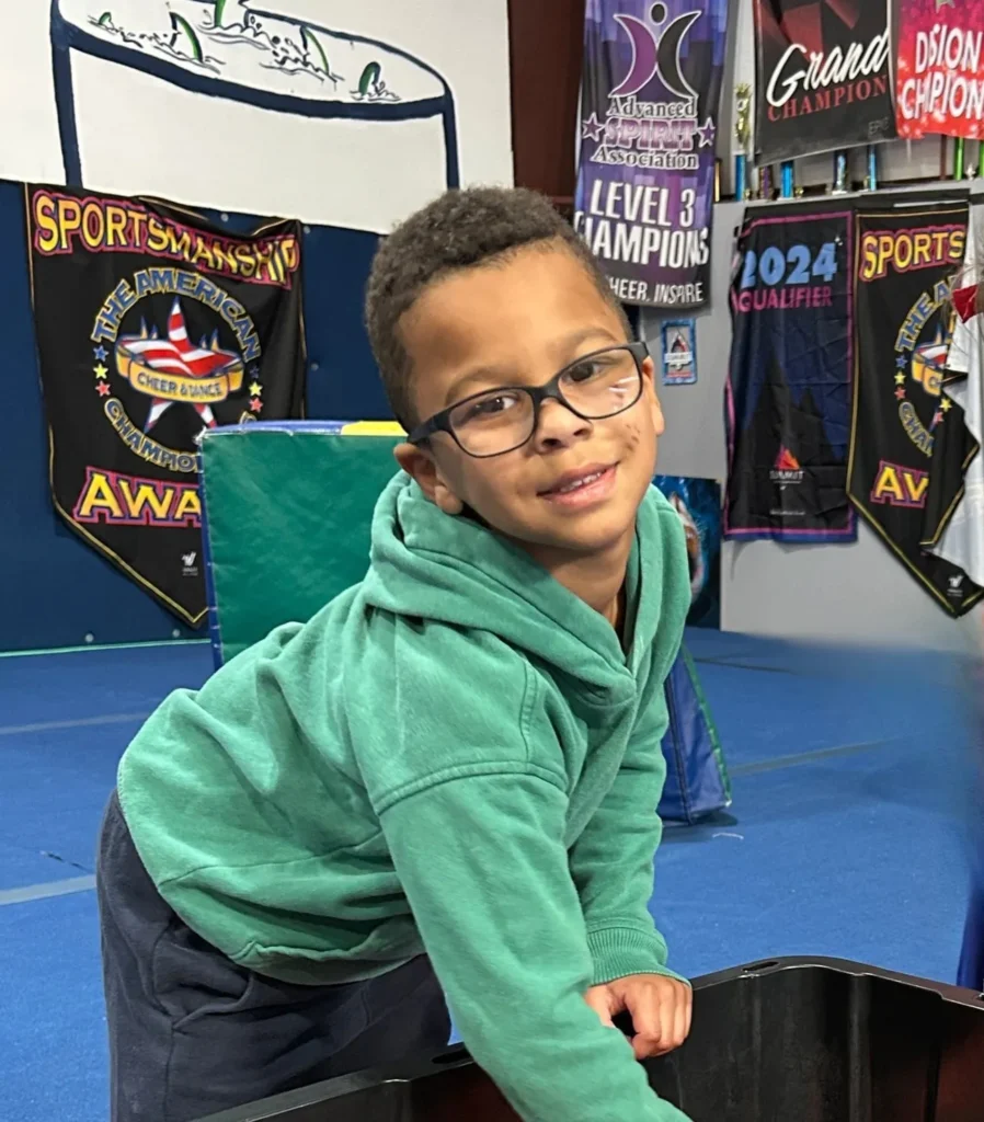 A smiling young boy wearing glasses and a green hooded sweatshirt leans forward in a gym setting with cheerleading banners in the background.